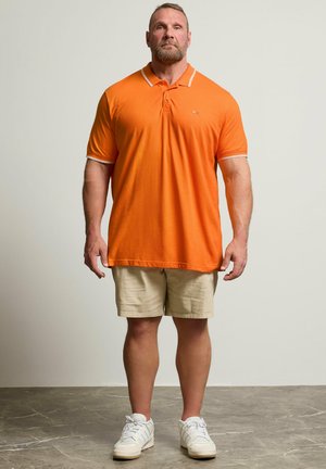 Hombre musculoso de pie mirando hacia adelante, vestido con una camisa polo naranja, pantalones cortos beige y zapatillas blancas sobre un fondo liso.