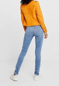 Semišová oranžová bunda s krátkým designem, v kombinaci s světle modrými skinny džíny a bílými teniskami, viděno zezadu.