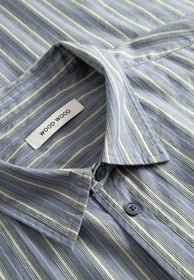 Srajca s sponko iz svetlo modrega blaga z navpičnimi črtami v beli in temnejši modri barvi. Ima ovratnik in dva temna gumba. Oznaka: WOOD WOOD.