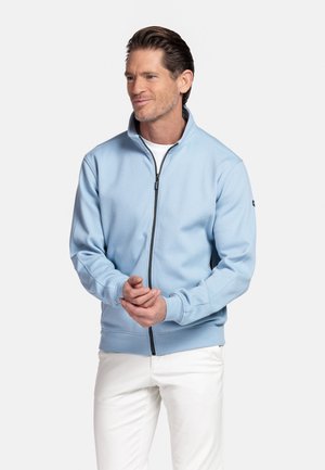 Man draagt een lichtblauwe jas met rits over een wit shirt en witte broek, staat met handen samengevouwen en kijkt iets naar de zijkant.