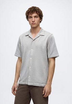 Jeune homme aux cheveux bouclés portant une chemise à manches courtes gris clair avec boutons et un pantalon marron, debout devant un fond blanc uni.