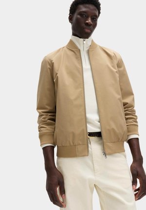 Junger Mann, der eine beige Bomberjacke über einem weißen Pullover mit Reißverschluss am Kragen und cremefarbigen Hosen trägt und vor einem einfarbigen Hintergrund steht.