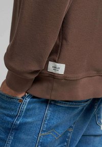 Sudadera marrón con textura acanalada, con una etiqueta de logo que dice "REPLAY" en el dobladillo, combinada con vaqueros de mezclilla azul.