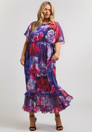 Frau trägt ein fließendes Maxikleid mit Blumenmuster in Lila-, Rosa- und Rottönen, hält den Rock und steht vor einem schlichten hellen Hintergrund.