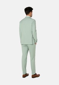 Hellgrüner Anzug mit taillierter Passform, strukturiertem Stoff und Schalkrippen. Kombiniert mit braunen Lederschuhen, von hinten betrachtet.