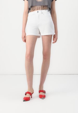 Person iført hvide shorts med opviklede kanter, grå cropped top med frynser og røde højhælede sko står mod en hvid baggrund.