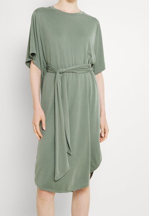Mujer que lleva un vestido verde oliva de corte holgado, hasta la rodilla, con mangas cortas y un cinturón de tela atado en la cintura.