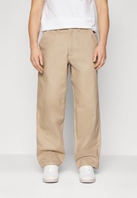 Beige vida byxor i bomull med synlig sömnad och en avslappnad passform. Bärs med vita sneakers, utan märkvärdiga mönster.