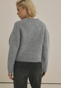 Grå stickad tröja med ribbad nederkant och axeldetalj, bärs tillsammans med svarta denimjeans, uppställd mot en enkel bakgrund.