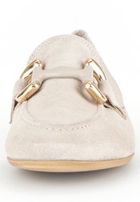 Beige suede slip-on shoe with a rounded toe, featuring a gold-tone accent and strap detail across the front. Flat sole visible.