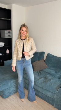 Tânără purtând o jachetă bej matlasată, bluză neagră și blugi evazați albaștri, stând lângă o canapea turcoaz într-un living minimalist.