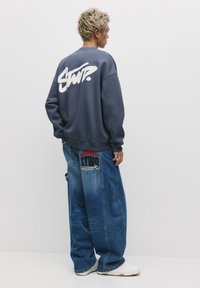 Tengerészkék pulóver nagy fehér grafikus szöveggel a hátsó részen, kiegészítve oversized kék farmer cargo nadrággal, melyen folt részletek találhatók.