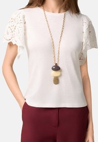 Top bianco a maniche corte con dettagli in pizzo, abbinato a una lunga collana d'oro con pietre ovali impilate di vari colori.