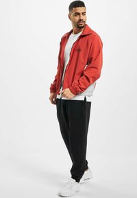 Jachetă roșie ușoară cu închidere prin fermoar, margini gri în contrast și logo brodat. Asortată cu pantaloni jogger negri și adidași albi.