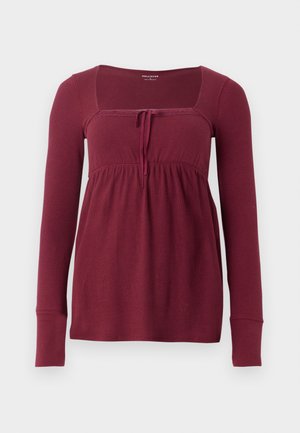 Pika pikkade varrukatega burgundi värvi topp, millel on ruudukujuline kaelus, pitsaktsendid ja sõlme detail rinna all, lõdva lõike ja pehme tekstuuriga.