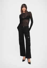 Top lungo in pizzo nero a maniche lunghe con motivo ondulato; pantaloni cargo neri a vita alta con tasche frontali e dettagli in cerniera argentata; décolleté neri a punta.