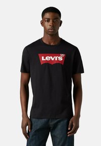 Joven vistiendo una camiseta negra de Levi's con un logo rojo y blanco, combinada con jeans azul oscuro, de pie contra un fondo liso.