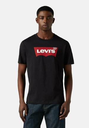 Ung mand iført en sort Levi's t-shirt med et rødt og hvidt logo, parret med mørkeblå jeans, stående foran en ensfarvet baggrund.