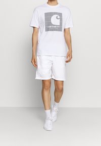 Bílá bavlněná tričko s šedým grafickým logem, spárované s bílými cargo šortkami. Nosené s bílými teniskami a kotníkovými ponožkami. Jednoduchý, ležérní outfit.