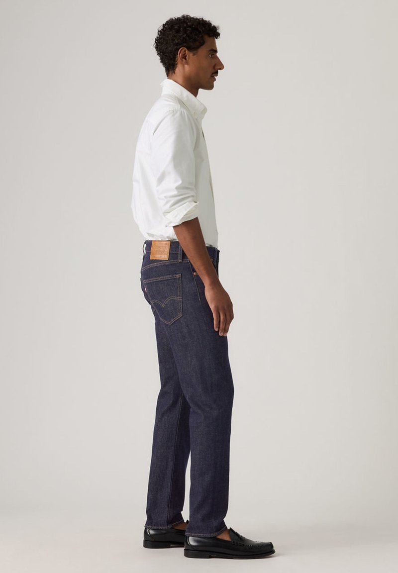 Hombre de perfil, vestido con camisa blanca de manga larga, vaqueros azul oscuro y zapatos de vestir negros, sobre un fondo claro y liso.