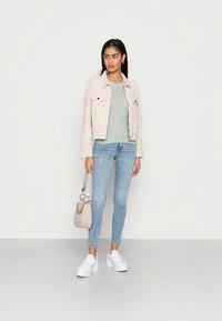 Casaco bege cropped com dois bolsos frontais, camisola verde clara, jeans skinny azul claro desgastados e ténis plataforma brancos. Mala pequena cinza.