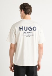 Cremfarbentes Baumwoll-T-Shirt mit lockerem Schnitt. Mit auffälligem, navy-blauen Text "HUGO" und verschiedenen Details auf der Rückseite, einschließlich "MCMXCIII."