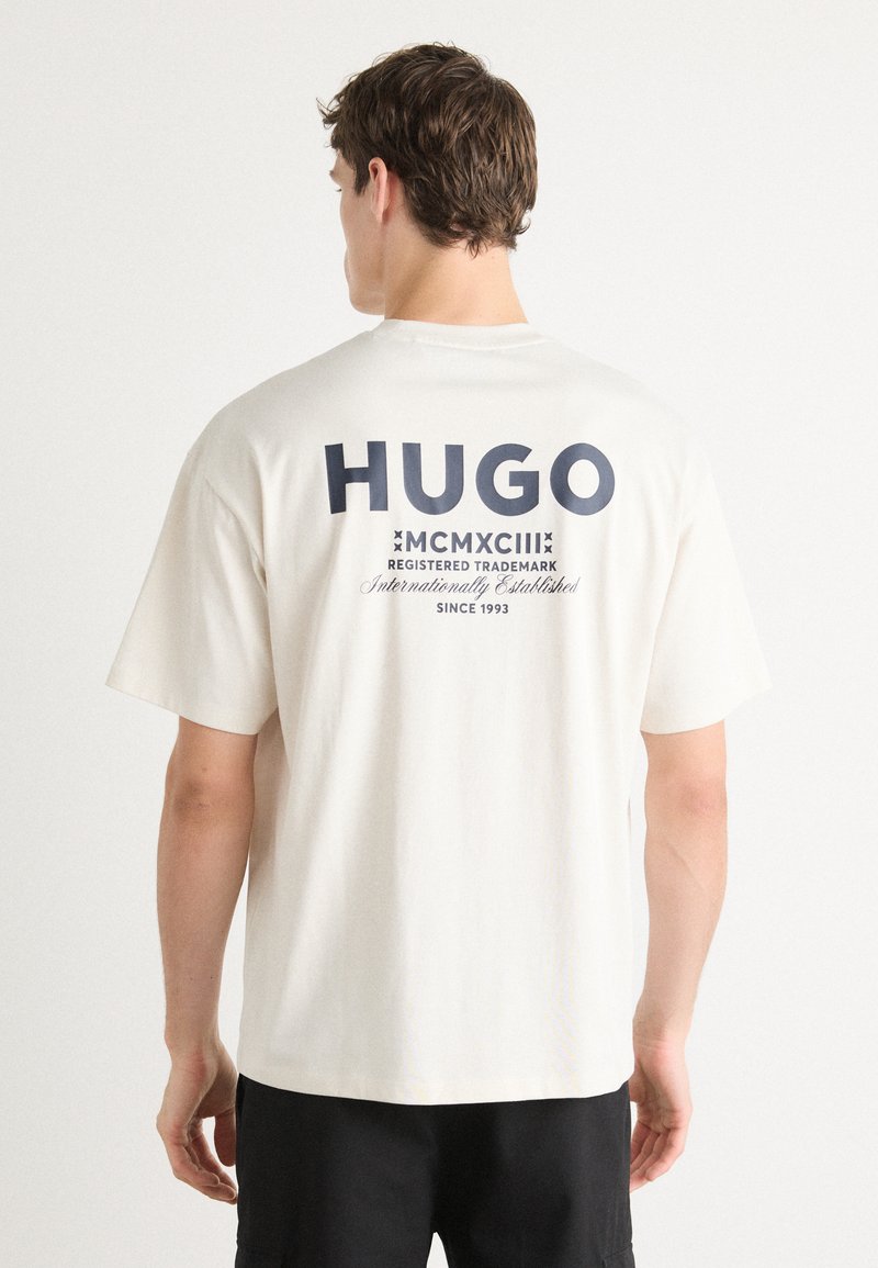 Cremfarbentes Baumwoll-T-Shirt mit lockerem Schnitt. Mit auffälligem, navy-blauen Text "HUGO" und verschiedenen Details auf der Rückseite, einschließlich "MCMXCIII."