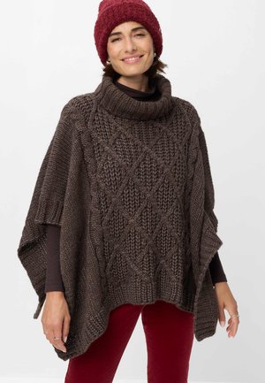 Kvindelig smilende iført en mørkebrun chunky strikket poncho, rød strikket hue og røde bukser mod en ensfarvet hvid baggrund.