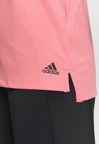 Chemise de sport rose avec fentes latérales, en tissu lisse. Présente un logo adidas noir sur l'ourlet inférieur. Portée avec des leggings noirs.