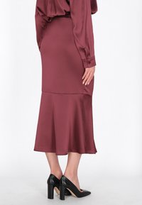 Burgundská saténová sukně s rozšířeným lemem, disponující hladkou texturou a jemným leskem. Kombinováno s černými botami na vysokém podpatku.