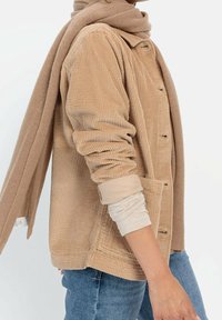 Veste en velours côtelé beige avec une texture côtelée visible, plusieurs boutons et deux poches avant. Superposée avec une écharpe marron clair.