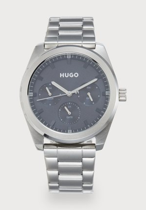 Montre quartz avec un cadran gris, bracelet en acier inoxydable argenté, dotée d'indicateurs de date et de jour, ainsi que de trois sous-cadrans pour les heures, la date et un affichage 24 heures.