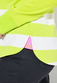 Maglione a righe verdi e bianche con orlo curvo, caratterizzato da dettagli rosa al taglio laterale e da un piccolo logo infinito giallo.