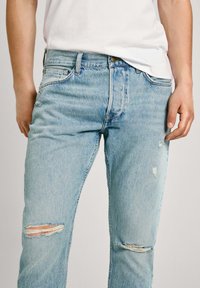 Jeans de mezclilla azul claro con un corte relajado, que presentan desgastes en las rodillas, diseño clásico de cinco bolsillos y cierre de botón.