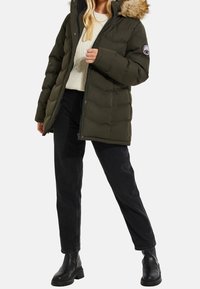 Femme portant une doudoune d'hiver verte avec capuche en fourrure, un pull en tricot crème, un jean noir et des bottines noires, debout sur un fond blanc.