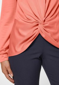 Perzikkleurig top met een twistdetail bij de taille, gecombineerd met donkerblauwe broek. De top heeft lange mouwen en een zachte, gladde textuur.