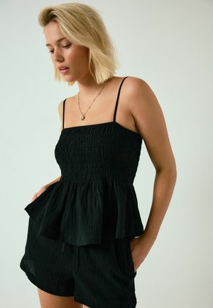 Blonde woman wearing a black textured sleeveless top and matching shorts with hands in pockets, looking downward, against a plain background.