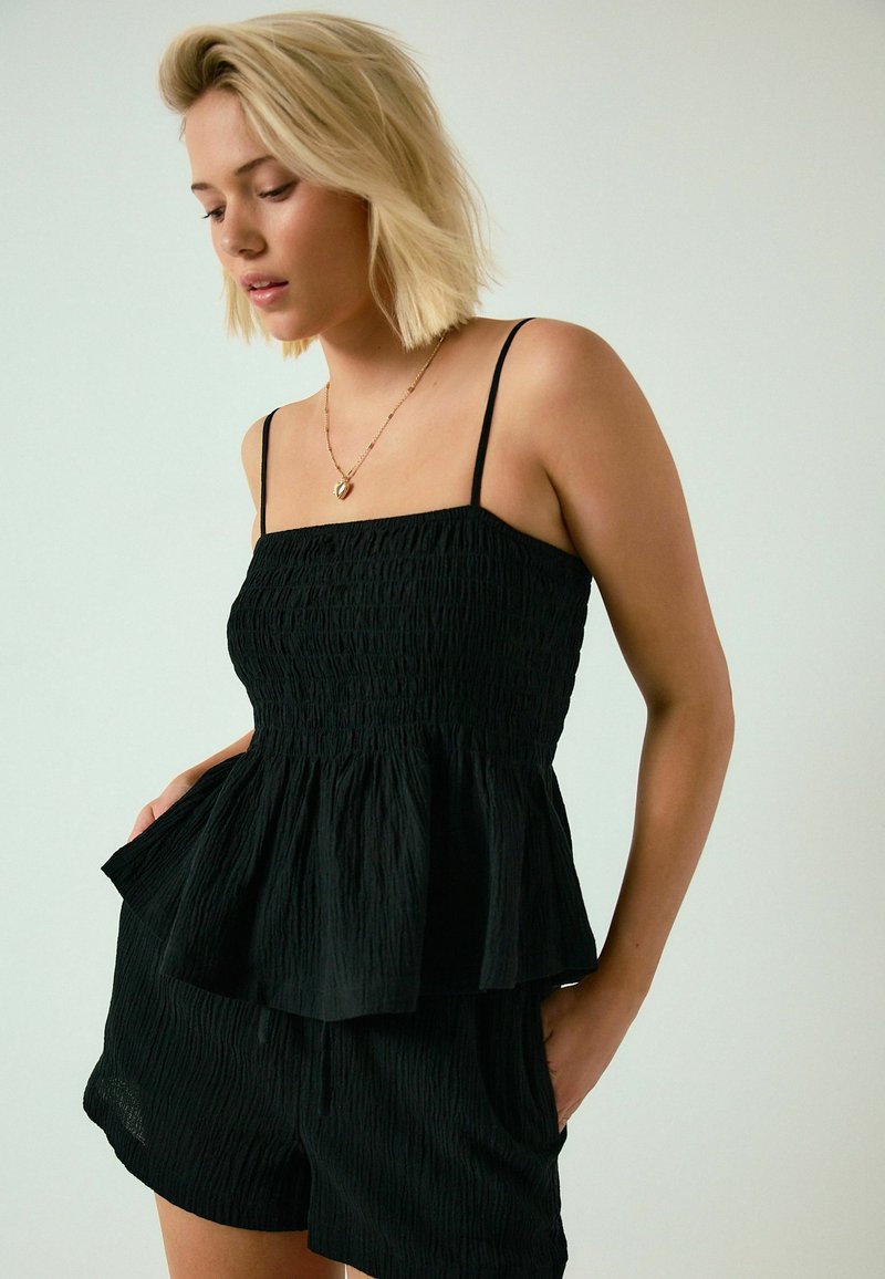 Mujer rubia con top sin mangas negro texturizado y shorts a juego, con las manos en los bolsillos, mirando hacia abajo, sobre un fondo liso.