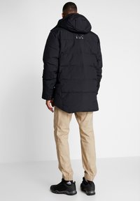 Black puffer jacket with a hood, featuring a smooth texture and a subtle logo on the back. Worn with beige pants and black sneakers.