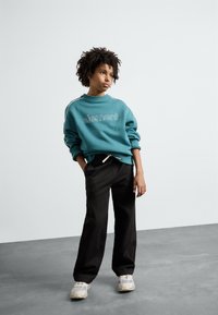 Sweatshirt teal avec logo et rayures blanches ; pantalon noir ample ; baskets grises ; tissu texturé ; coupe décontractée.