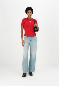 T-shirt rouge à manches courtes avec un logo circulaire, associé à un jean large bleu clair et des bottines noires. Le mannequin tient un sac à main noir.
