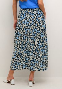 Kvinna iförd en svart blommig midi-skjol med blå och beige blommor, parat med en blå topp och vita blockklackade mules.