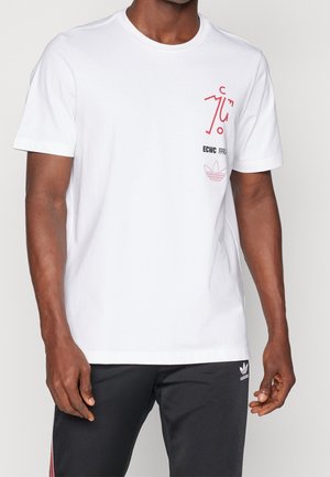 Man wearing a white T-shirt with red abstract print and black text on chest, paired with black Adidas pants with a white logo.