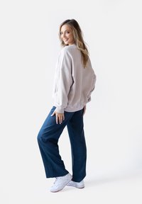 Jasnoróżowa, oversize'owa bluza z prążkowanym wykończeniem, w połączeniu z ciemnoniebieskimi spodniami o szerokich nogawkach i białymi trampkami na jednolitym tle.