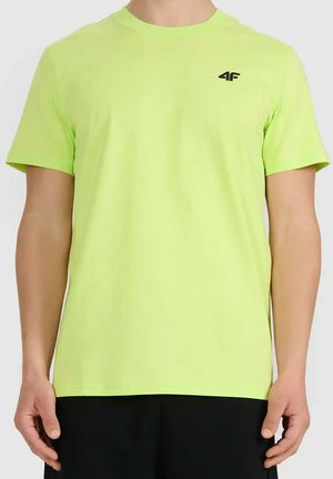 Mężczyzna w neonowo zielonej koszulce z krótkim rękawem z małym czarnym logo "4F" na piersi, w połączeniu z czarnymi spodniami na jednolitym tle.