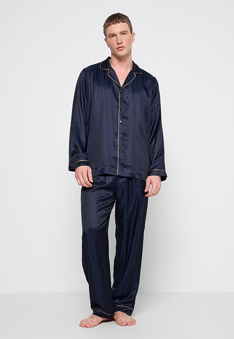 Jeune homme debout pieds nus, portant un pyjama en satin bleu marine avec des bordures dorées sur fond blanc uni.