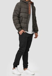 Homme portant une veste rembourrée vert foncé avec une doublure orange, un t-shirt blanc, un pantalon de jogging noir et des baskets blanches, se tenant avec une main dans la poche.