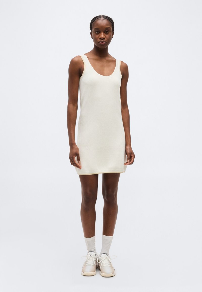 Γυναίκα που στέκεται φορώντας αμάνικο, μπεζ φόρεμα μέχρι το γόνατο με λευκές κάλτσες και μπεζ αθλητικά παπούτσια, μπροστά σε απλό φως φόντο.