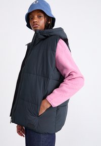Gilet imbottito nero con cappuccio, dotato di chiusura con zip e tasche laterali. Indossato sopra una maglietta a maniche lunghe in pile rosa. Cappello a secchiello di maglia blu.