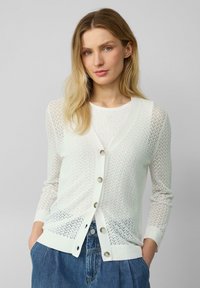 Leichter weißer Cardigan mit Zickzackmuster, der einen tiefen V-Ausschnitt, vordere Knöpfe und langen Ärmeln hat. Getragen über einem strukturierten Oberteil.